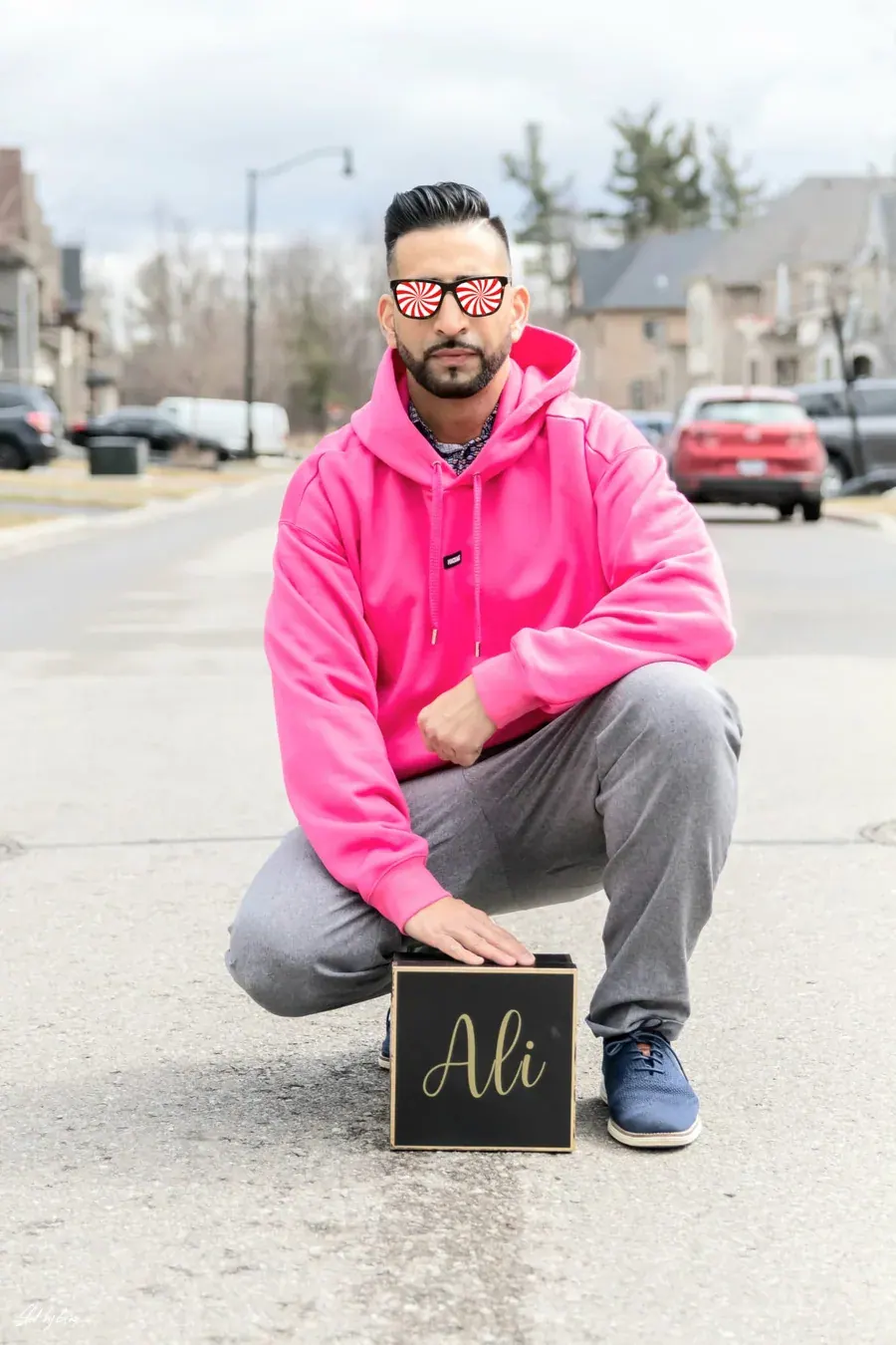 Ali crouched in the middle of a suburban street in a pink hoodie and spiral sunglasses, one hand on a black Ali box